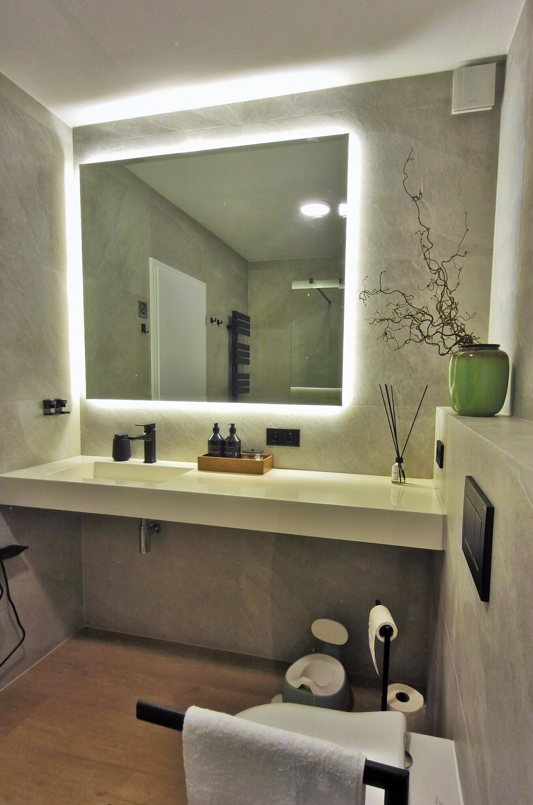 A modern bathroom with a large mirror, a sink with a wooden tray, a green vase, and a toilet with a towel.
