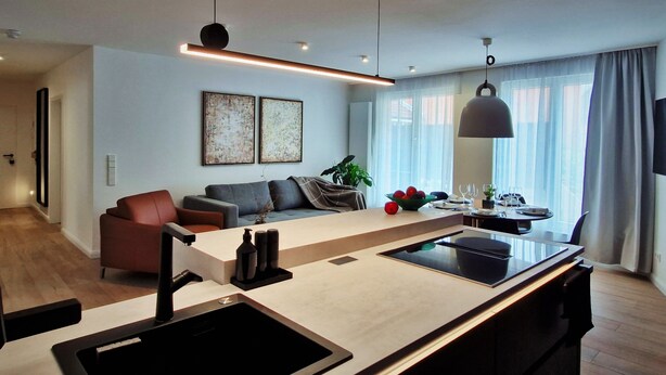 A modern kitchen with a central island, a dining area with a table and chairs, a sofa, and a large window with curtains.