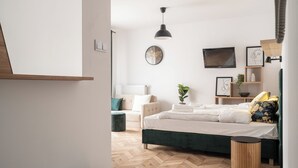 Een moderne slaapkamer met een bed, een rond houten bijzettafeltje, een zwarte hanglamp, een televisie en een wandplank met decoratieve objecten.