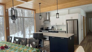 A modern kitchen with a central island, white cabinets, stainless steel appliances, and a dining area with a table and chairs. - Lake, Spa & Game Room | Le Havre du Voyageur (La Minerve)