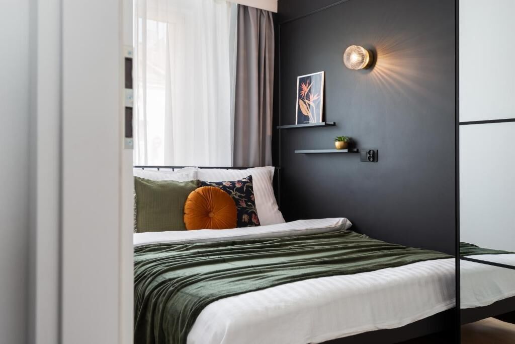 A modern bedroom with a bed, a window with curtains, a wall-mounted shelf with a plant, and a wall light.