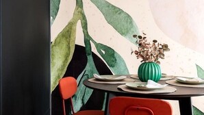 Een eethoek met een tafel gedekt voor twee personen, een groene vaas met gedroogde bloemen en een groot abstract blad-muurschildering op de muur.