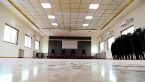 A spacious, well-lit conference room with a polished floor, a stage with chairs, and a projector screen.