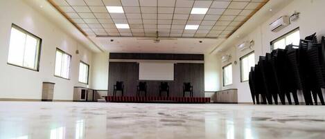 A spacious, well-lit conference room with a polished floor, a stage with chairs, and a projector screen.