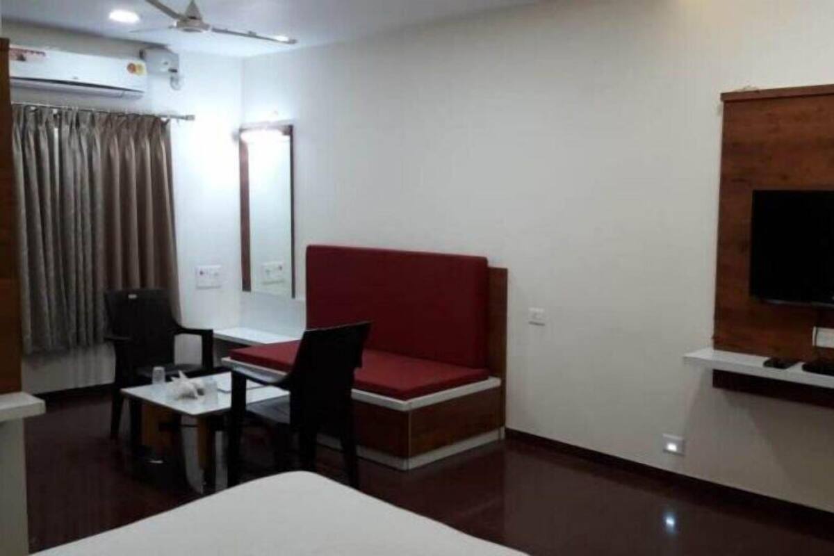 Habitación de hotel con sofá rojo, televisor montado en la pared, escritorio con silla y equipo de aire acondicionado.