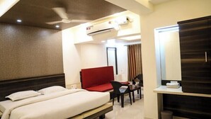 A hotel room with a bed, a red sofa, a desk with a chair, a mirror, and an air conditioning unit.