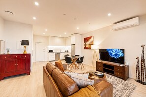 A modern living room with a brown leather sofa, a dining area with a table and chairs, a flat-screen TV, and a kitchen in the background.
