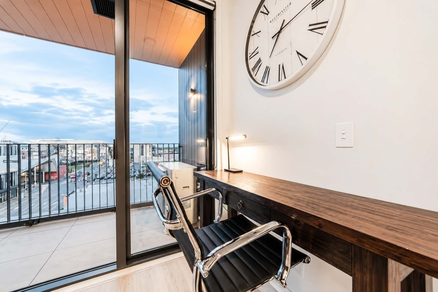 Una habitación moderna con un ventanal grande, un escritorio de madera, una silla y un reloj de pared.