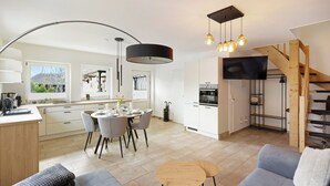A modern kitchen with white cabinets, a dining area with a round table and chairs, a grey sofa, and a staircase leading to a loft.