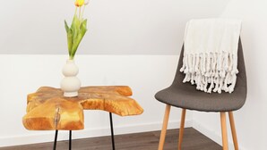 A room with a wooden side table, a vase with flowers, and a chair with a white throw blanket.