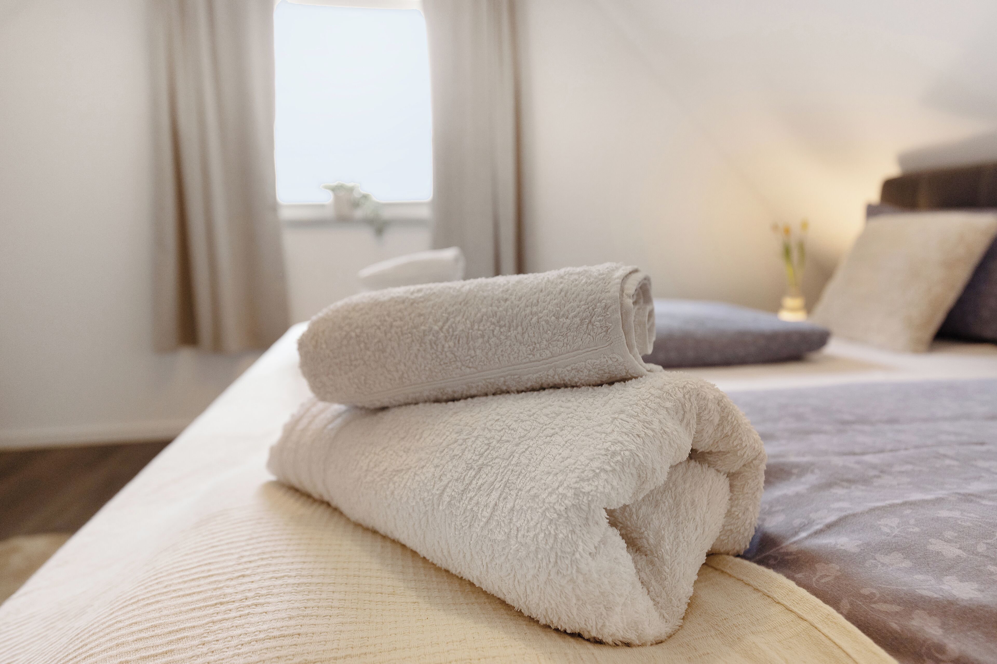 Een netjes opgemaakt bed met een opgevouwen beige handdoek erop, een grijs kussen en een witte vaas met bloemen op het nachtkastje.