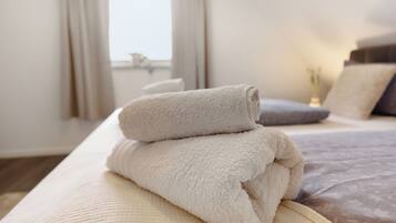 Een netjes opgemaakt bed met een opgevouwen beige handdoek erop, een grijs kussen en een witte vaas met bloemen op het nachtkastje.
