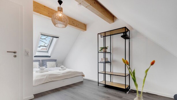 A bedroom with a skylight, a bed, a wooden floor, a black metal shelving unit, and a vase with flowers.