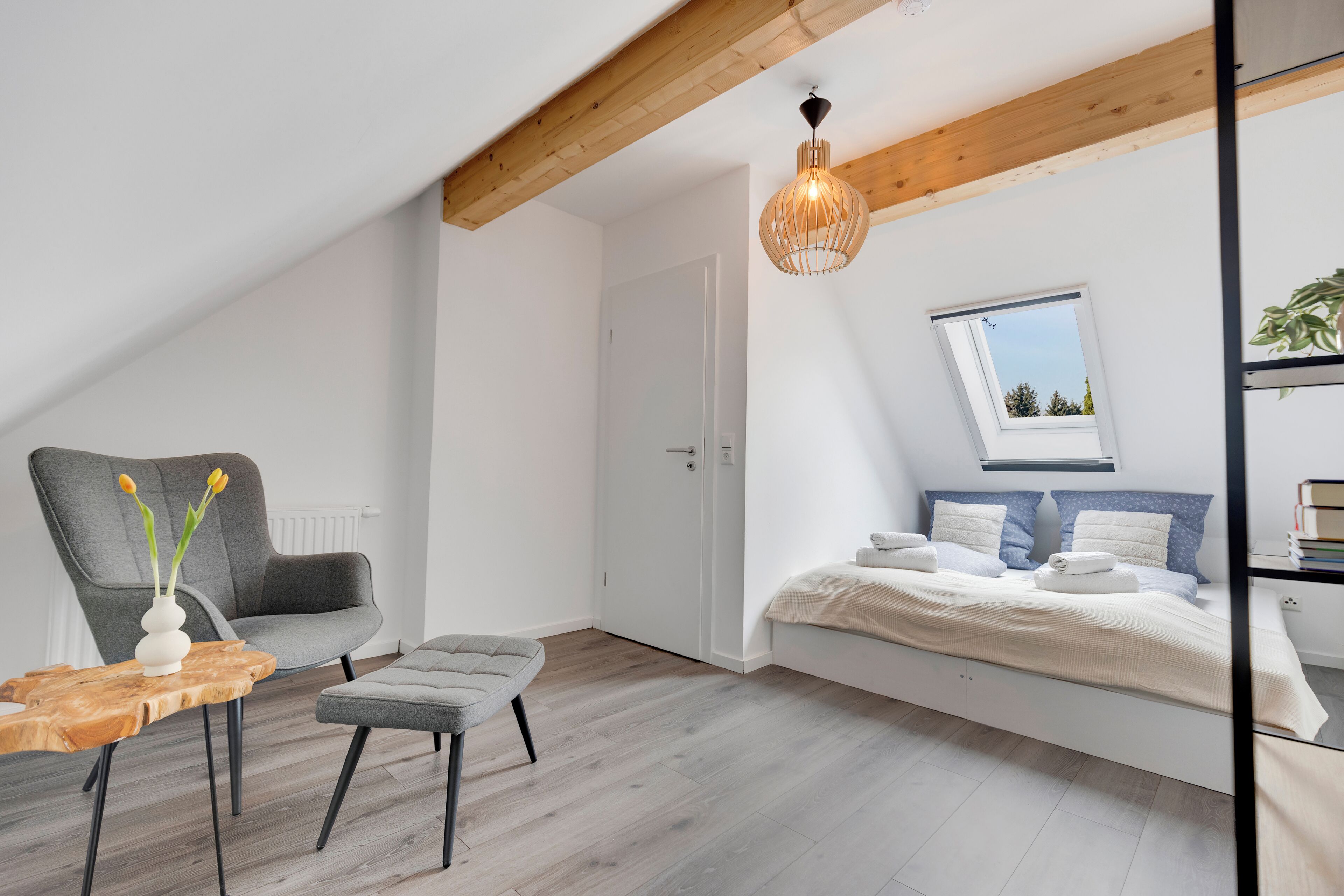 Una camera da letto moderna con soffitto spiovente, un letto con biancheria da letto bianca, una poltrona grigia, un piccolo tavolino e una lampada a sospensione.