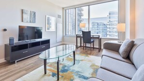 Una sala moderna con una mesa de centro de vidrio, un sofá, un televisor en un mueble negro y un amplio ventanal con vistas a la ciudad.