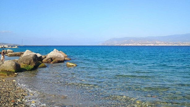 A coastal scene with clear blue water, large rocks, and a distant cityscape. - Apartment 204 (Messina)