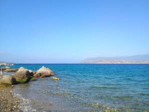 A coastal scene with clear blue water, large rocks, and a distant cityscape. - Apartment 204 (Messina)