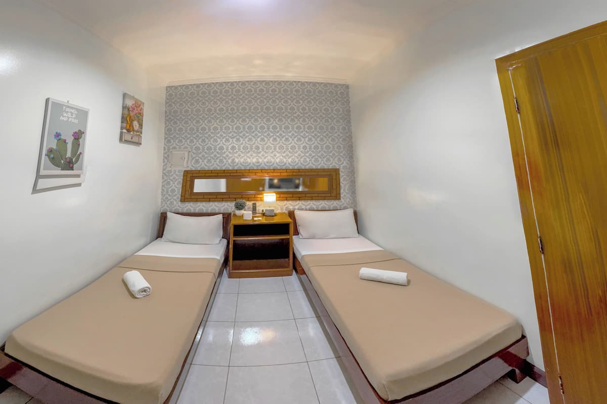 A small, narrow hotel room with two single beds, a wooden nightstand with a lamp, a small potted plant, and a framed picture on the wall.