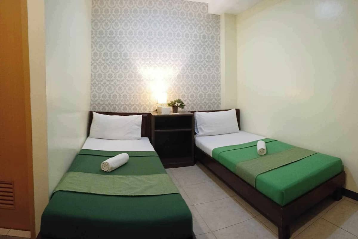 A small, clean room with two single beds, each with a green bedspread and white pillows. There is a small table with a lamp and a potted plant between the beds.