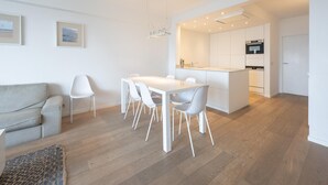 A modern kitchen with white cabinetry, a dining area with a white table and chairs, and a light-colored sofa in a spacious living area. - Spacious Seaside Apartment (Knokke-Heist)