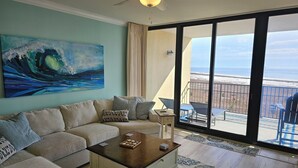 A living room with a beige sectional sofa, a wooden coffee table, and a large painting of a wave. There is a balcony with a view of the beach. - Holiday Isle SandDollar 713 (Dauphin Island)