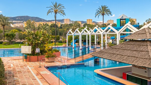 Una piscina con un puente, palmeras y vistas a edificios al fondo.