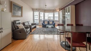 A modern living room with a sofa, a blue armchair, a dining table with chairs, and a kitchen area with cabinets. - Apartment in Knokke Near Zeedijk Beach (Knokke-Heist)