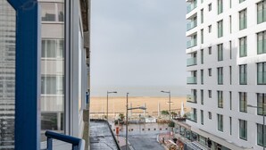 A view from a window overlooking a street with a beach in the distance, a building with balconies, and parked vehicles. - Apartment in Knokke Near Zeedijk Beach (Knokke-Heist)