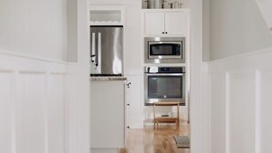 Un pasillo que conduce a una cocina con armarios blancos, un refrigerador de acero inoxidable, un microondas y un horno.
