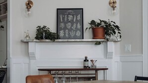 Un comedor con una mesa de madera, una chimenea, una ilustración enmarcada de un jardín de hierbas y plantas en macetas sobre la repisa.
