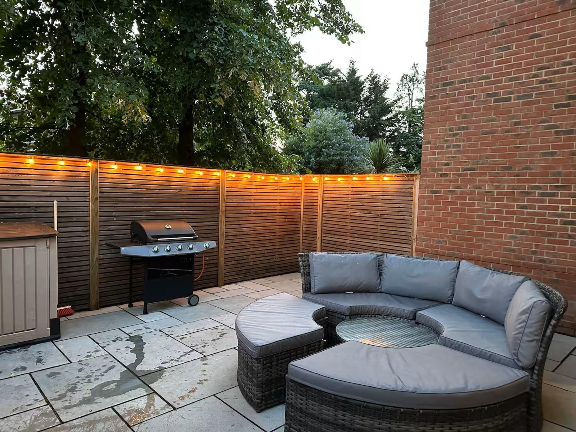 A patio with a curved grey outdoor sofa, a barbecue grill, and a wooden fence with string lights.