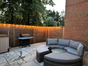A patio with a curved grey outdoor sofa, a barbecue grill, and a wooden fence with string lights. - Thames Ditton Apt - Sleeps 6 - Garden & Bbq (Thames Ditton)