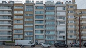 Une rangée d’immeubles résidentiels modernes à plusieurs étages, dotés de balcons, avec un fourgon blanc garé devant et quelques voitures stationnées le long de la rue.