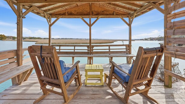A wooden deck with two rocking chairs and a small table, overlooking a calm body of water and distant shoreline. - Cedar Creek - Creek Front, Mainland, 3-bedroom (Pawleys Island)