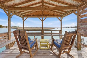 A wooden deck with two rocking chairs and a small table, overlooking a calm body of water and distant shoreline. - Cedar Creek - Creek Front, Mainland, 3-bedroom (Pawleys Island)