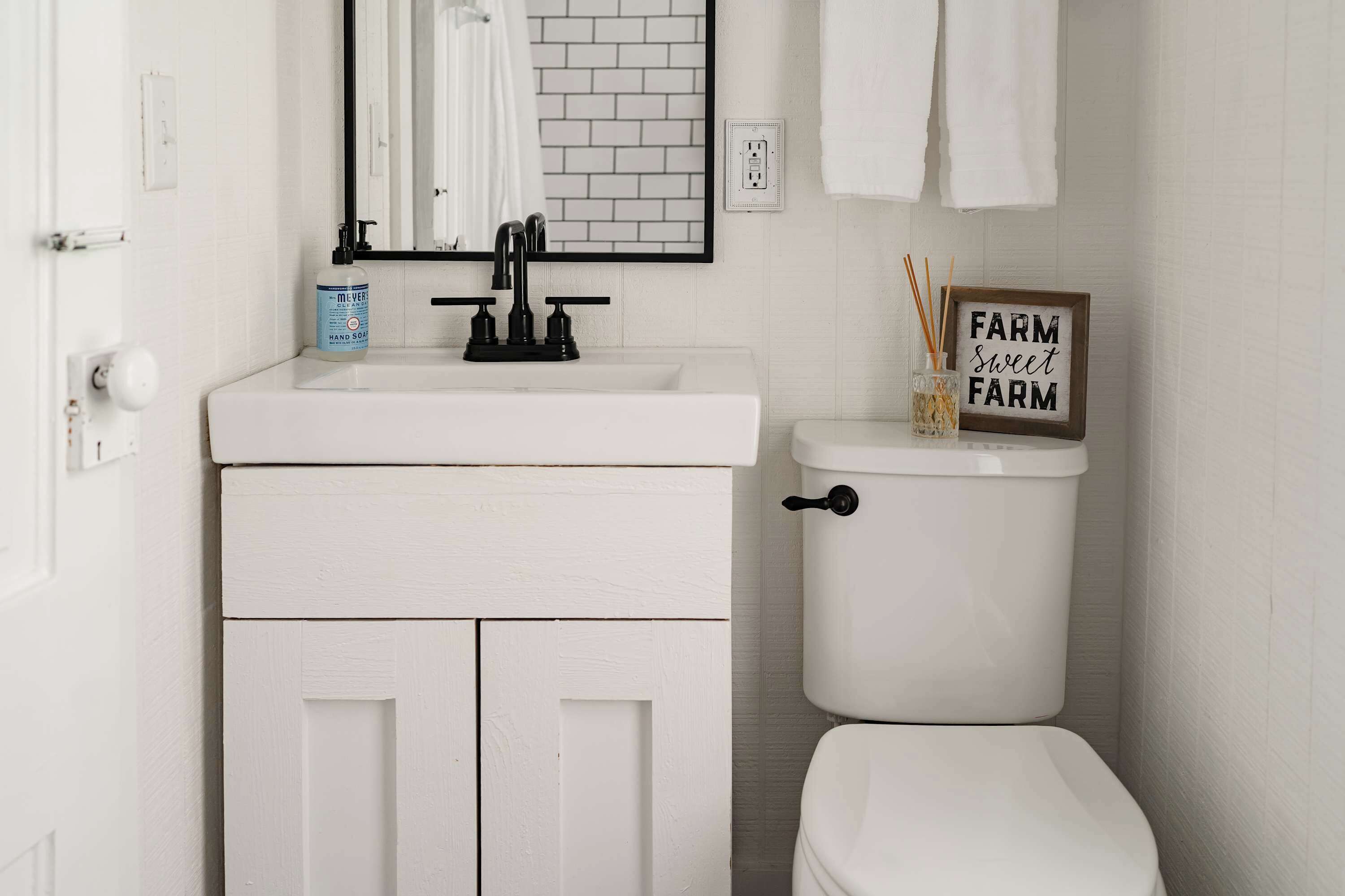 A white bathroom with a white sink, black faucet, and a toilet. There is a framed sign on the toilet that reads "FARM Sweet Farm".