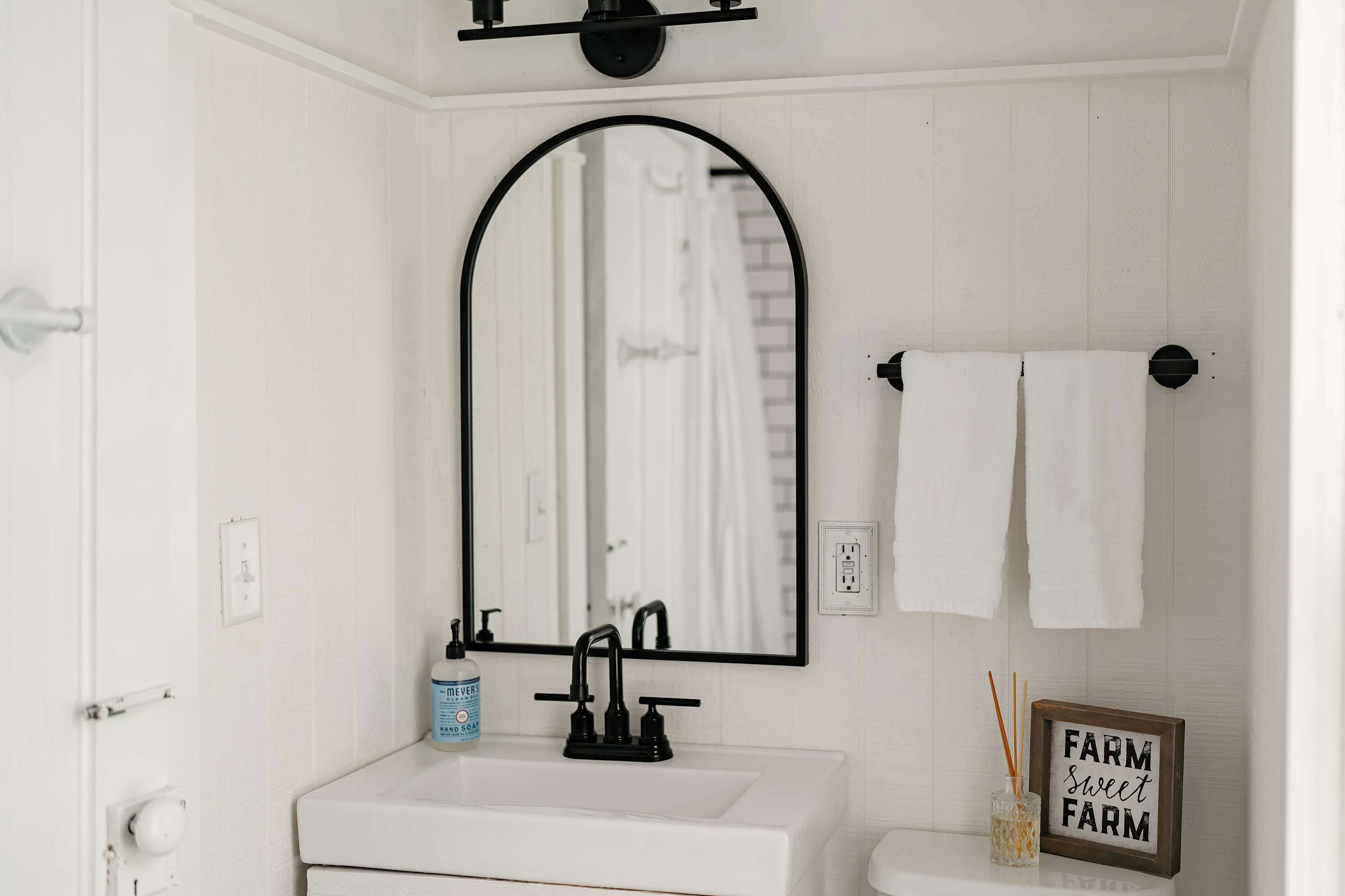Een badkamer met een witte wastafel, een zwarte kraan en een spiegel. Er is een handdoekhouder met twee handdoeken en een bord met de tekst 'FARM sweet FARM'.
