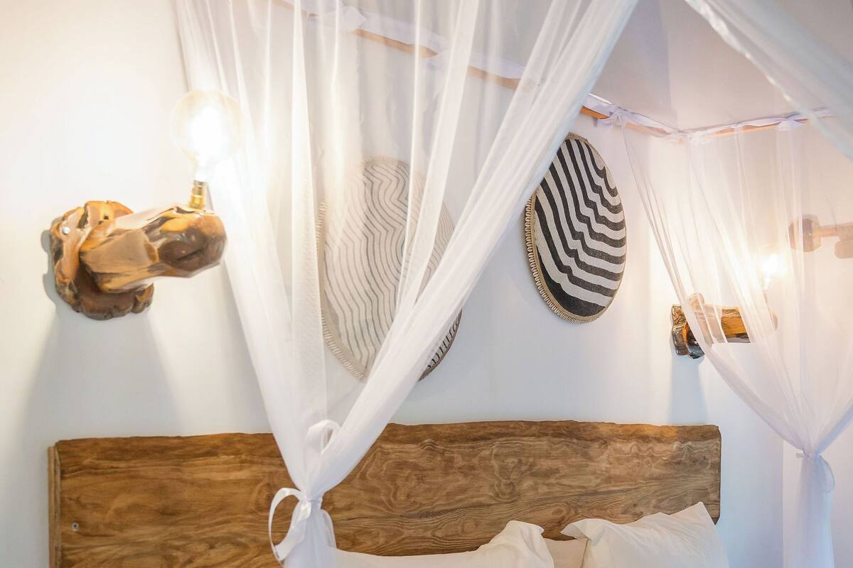 Una cama bien hecha con ropa de cama blanca, un cabecero de madera y un dosel blanco. Hay una mesita con un florero y una lámpara fijada a la pared.