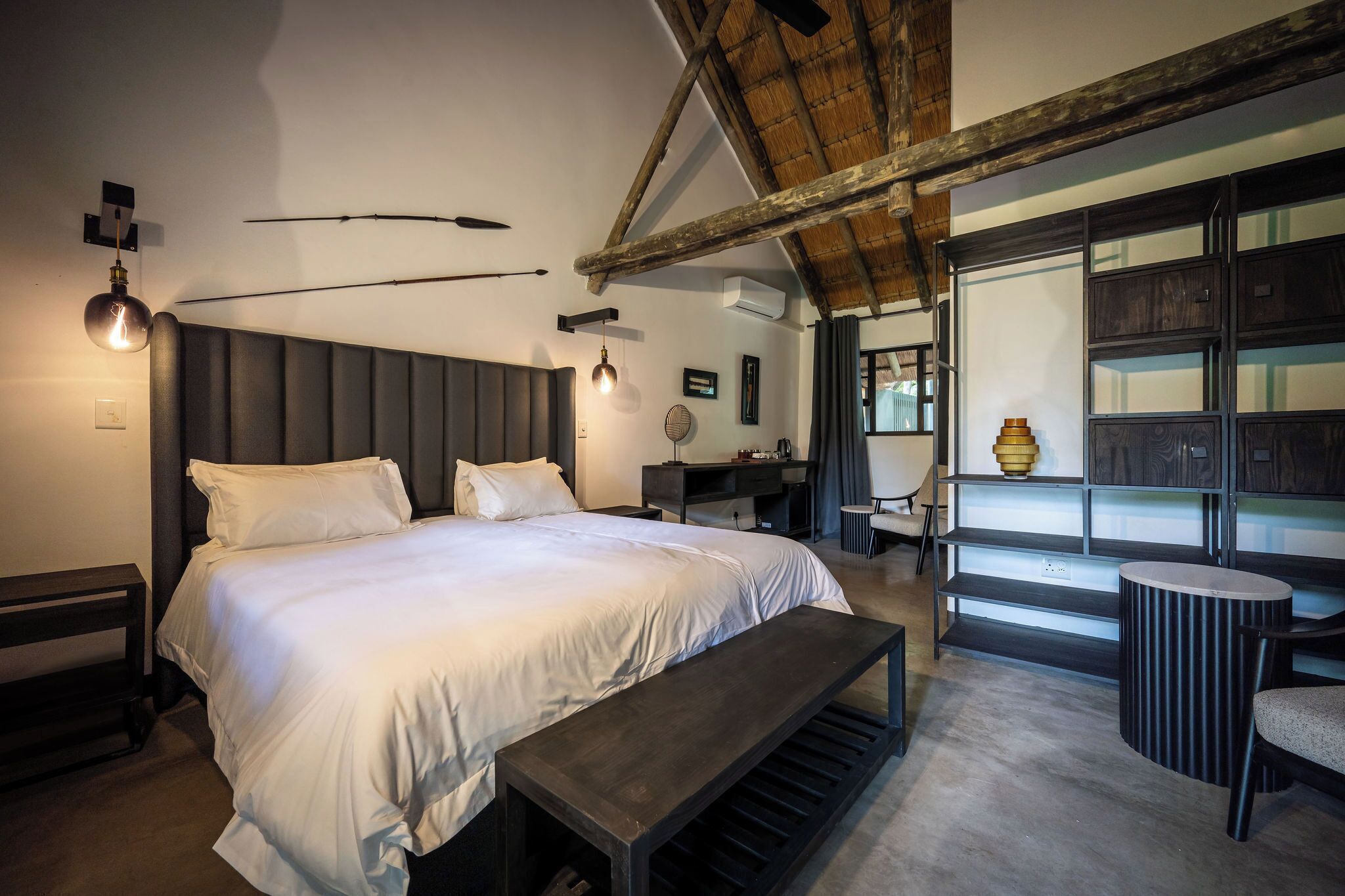A modern bedroom with a large bed, a bench, a desk, and a shelving unit.
