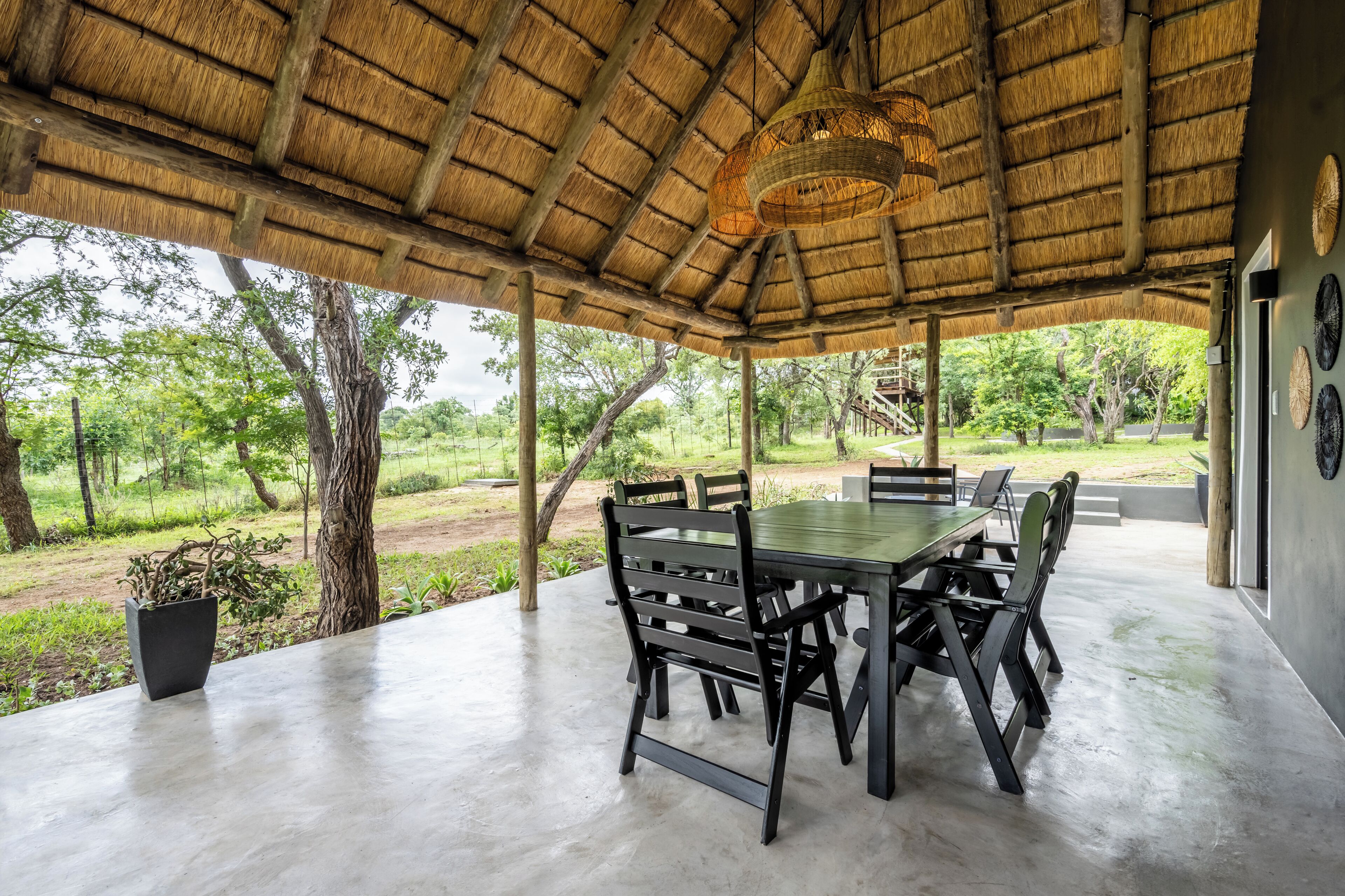 Un espacio de comedor al aire libre con techo de paja, una mesa con sillas y vistas al entorno verde.