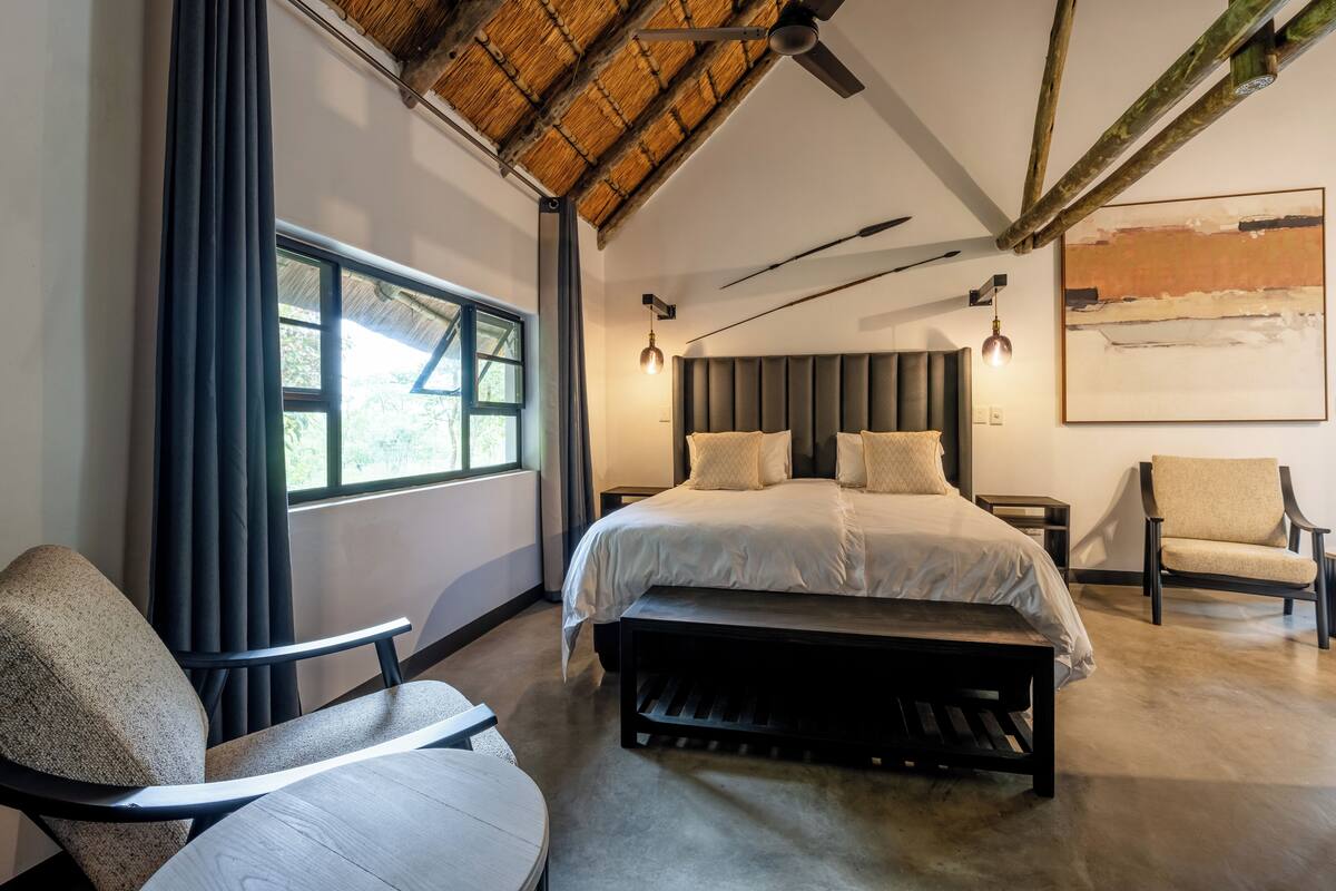 Un dormitorio moderno con una cama grande, un banco, dos sillas, un ventilador de techo y un ventanal amplio.