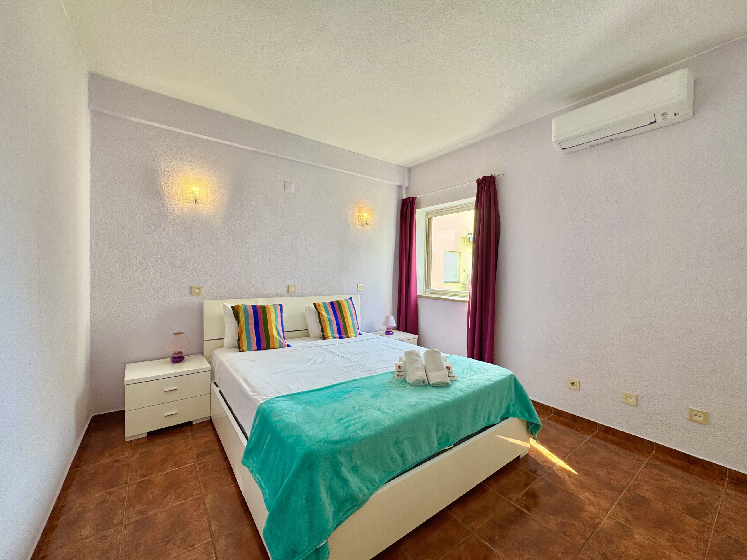 A bedroom with a bed, a nightstand, a window with curtains, and an air conditioning unit on the wall.