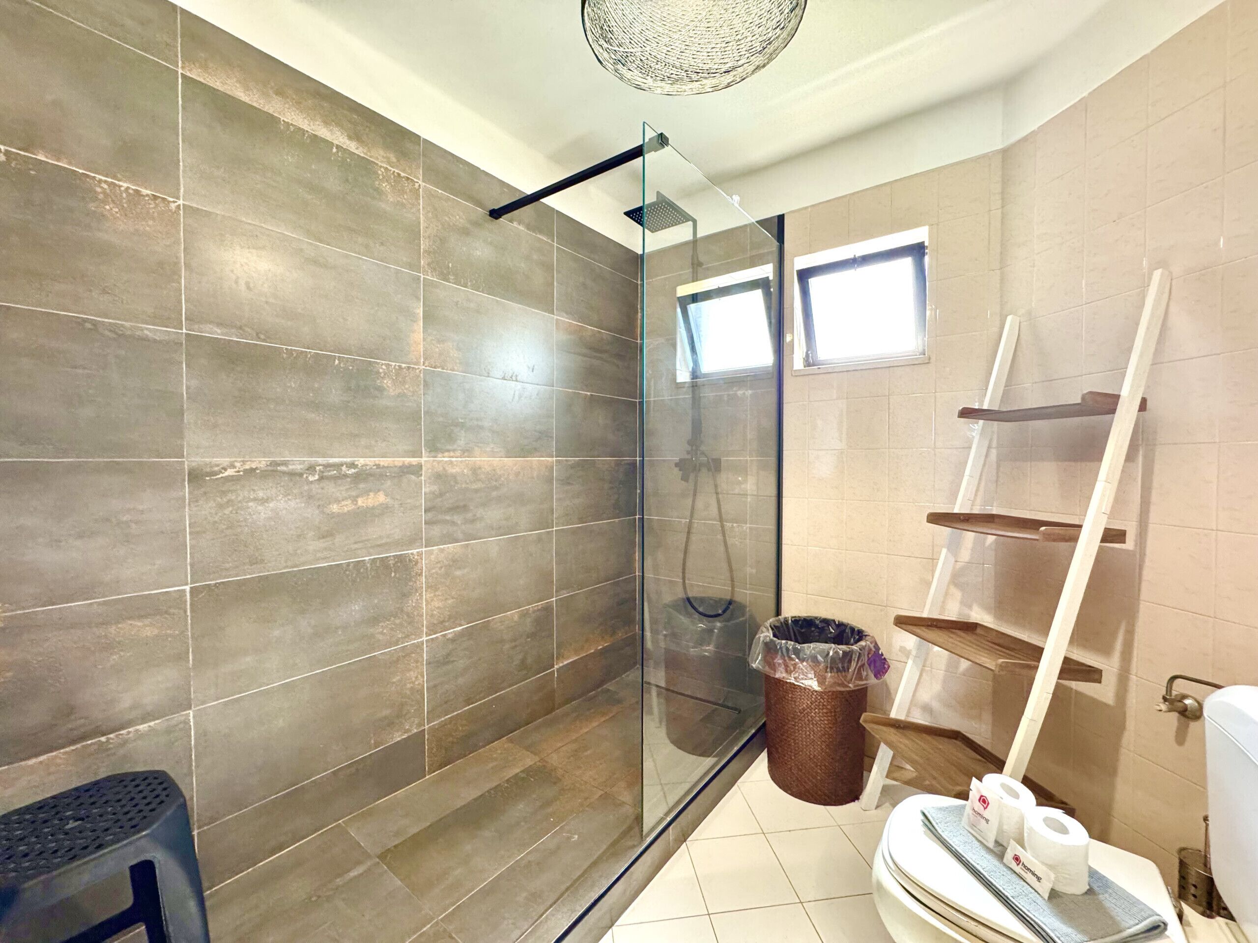 A modern bathroom with a glass-enclosed shower, a wooden ladder, a trash bin, and a toilet with toilet paper.
