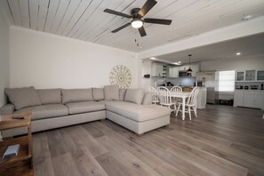 A modern living room with a sectional sofa, a dining area with white chairs, a kitchen with white cabinets, and a ceiling fan.