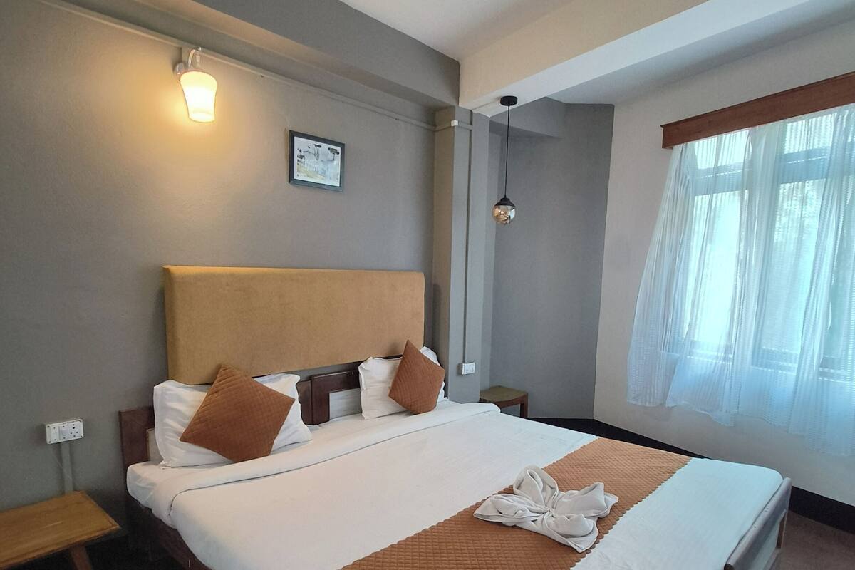 A hotel room with a large bed, a bedside table, a nightstand, a wall-mounted light, a window with sheer curtains, and a framed picture on the wall.
