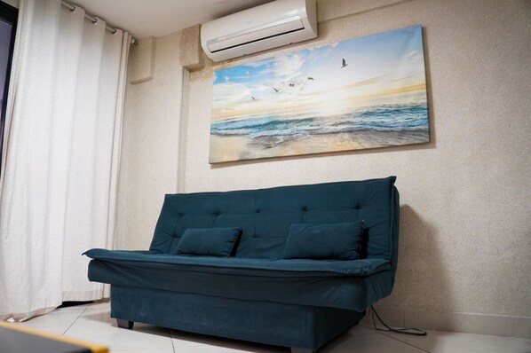 A teal sofa with two cushions in a room with a beach scene painting on the wall and an air conditioning unit above.