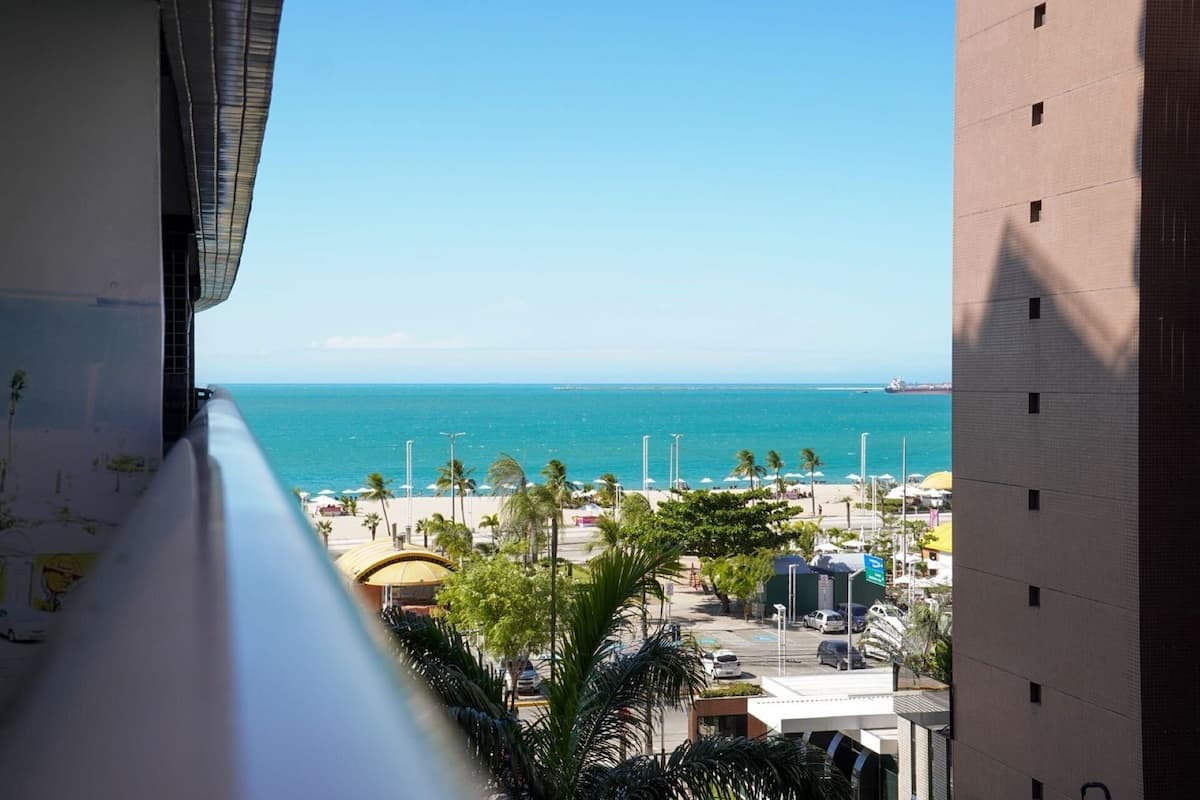 A view from a balcony overlooking a beach with a clear blue sea, palm trees, and a distant ship.