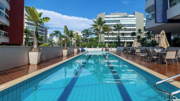 A swimming pool with lounge chairs and umbrellas, surrounded by palm trees and modern buildings. - Apto pé na areia na Riviera São Lourenço (Bertioga)