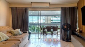 A living room with a sofa, a dining table with chairs, a fan, and a view of the city through large windows. - Apto pé na areia na Riviera São Lourenço (Bertioga)