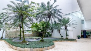A spacious indoor atrium with palm trees, a tiled floor, and a modern architectural design. - Apto pé na areia na Riviera São Lourenço (Bertioga)